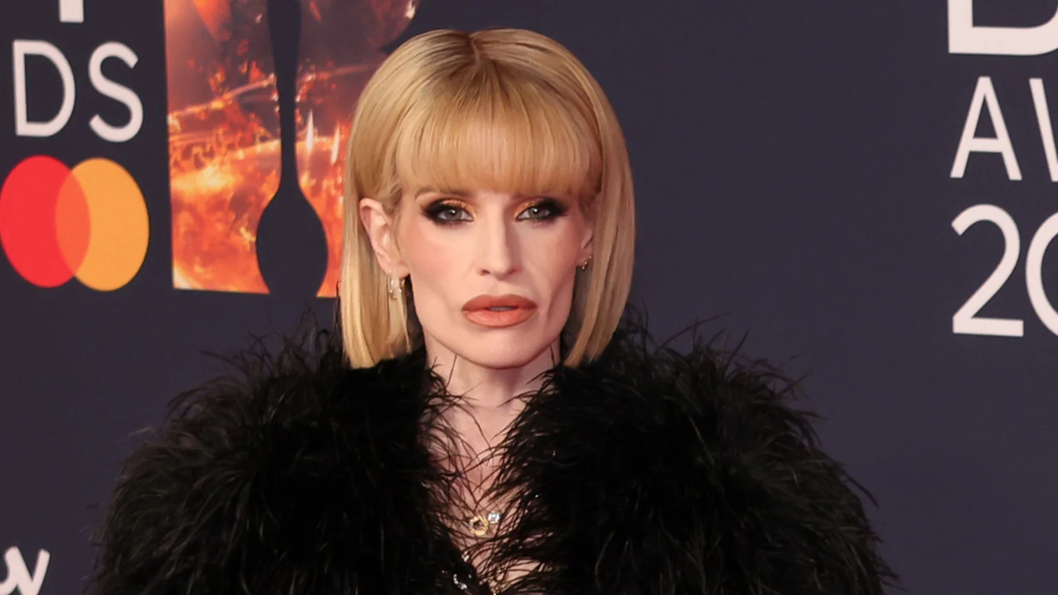 A person with blonde bob hair and bangs, wearing prominent eye makeup and light brown lipstick, sports a black feathered garment in front of a dark event backdrop.