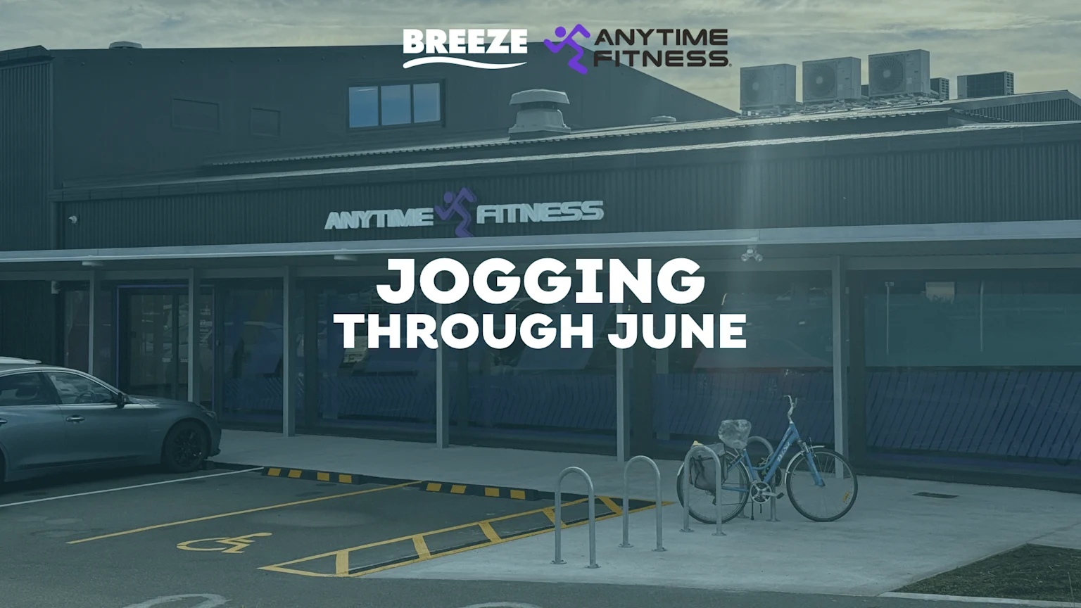 The exterior of an Anytime Fitness gym is visible with a prominent "Jogging Through June" text overlay, a parked car, and a bicycle at a bike rack.