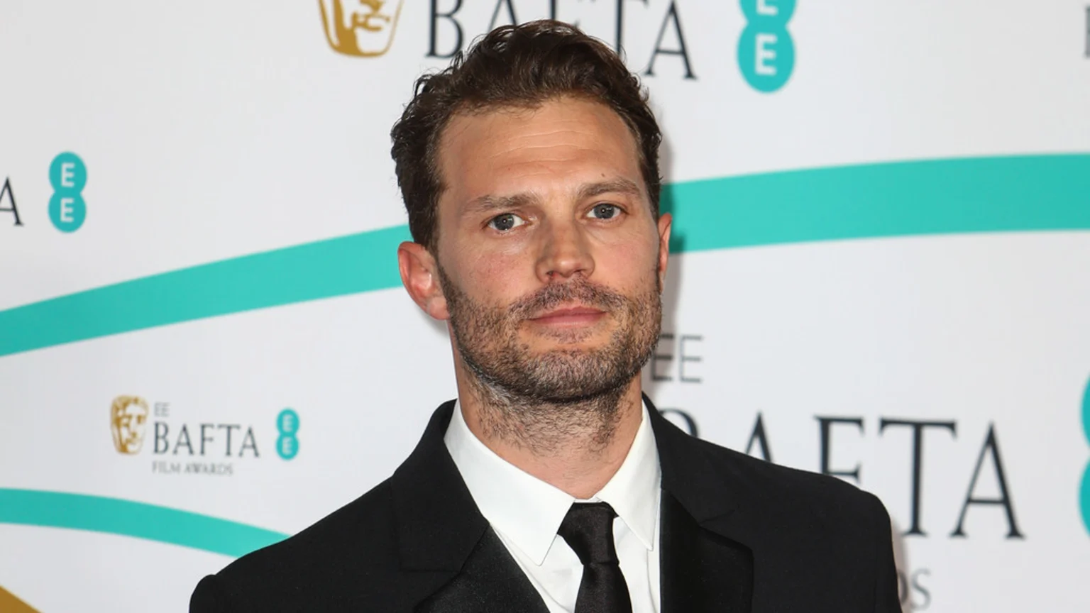 Jamie Dornan in a black suit and tie, with a beard, poses for a photo in front of a white background with BAFTA Film Awards logos.