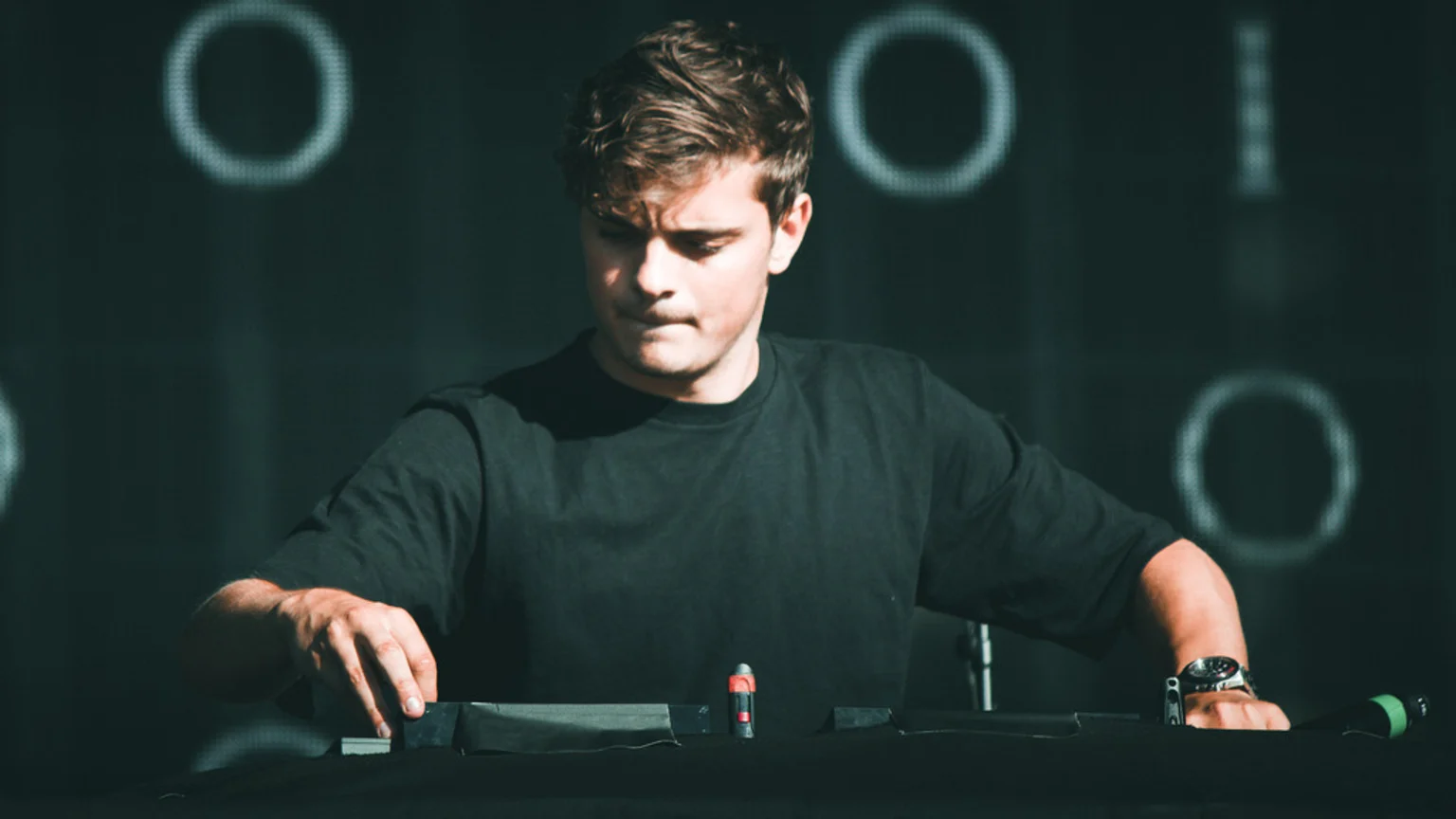 DJ Martin Garrix, wearing a black t-shirt, looks down intently at his mixing equipment with hands on the controls, against a dark background with blurred glowing circular shapes.