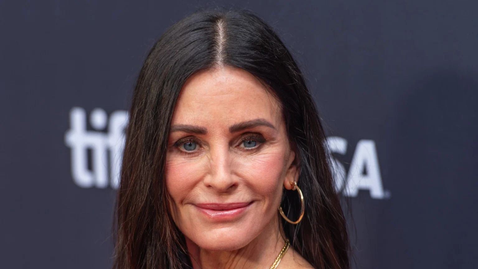 A close-up of Courteney Cox with dark hair parted in the middle, blue eyes, and light makeup, wearing gold hoop earrings and a gold necklace, against a blurred dark background.
