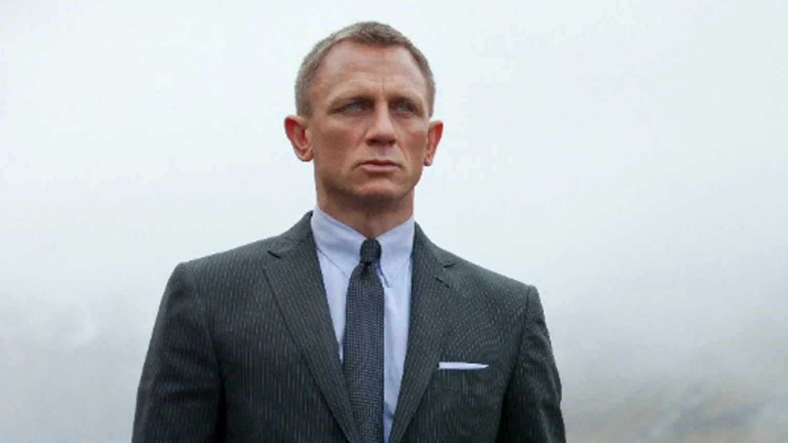 Actor Daniel Craig, portraying James Bond, in a pinstripe suit, light blue shirt, and dark tie, looks intently to the side against an overcast sky.