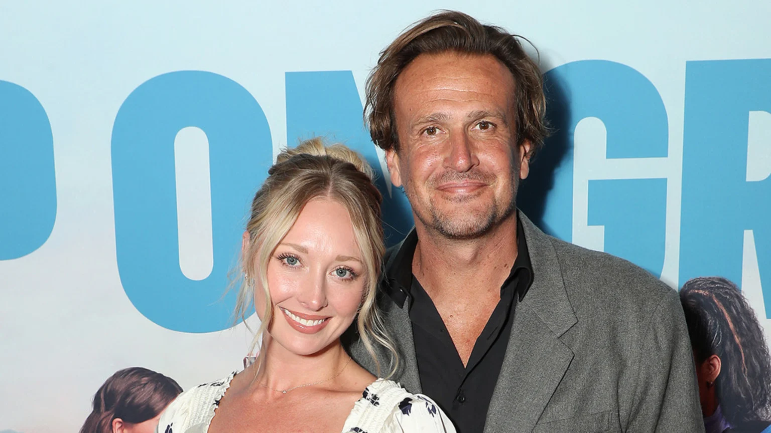 Jason Segel, smiling, stands next to a smiling blonde woman, both in front of a light blue background with partial text.