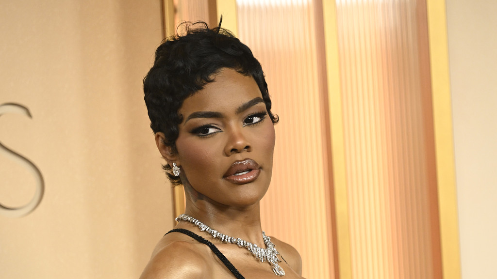 A woman with a dark, wavy pixie cut, winged eyeliner, and glossy lips wears diamond earrings and a necklace, looking toward the viewer against a warm, panelled background.