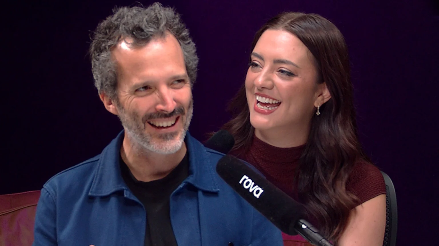 Bret McKenzie, a man with curly grey hair and beard, smiles, looking toward a laughing woman with long dark hair next to a microphone with 'rova' written on it.
