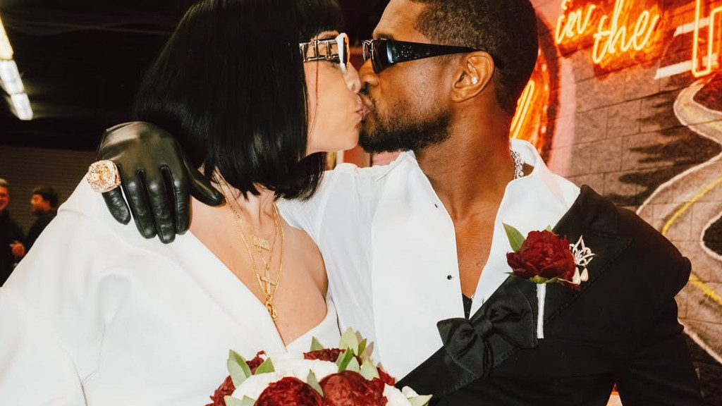 A woman with a black bob and sunglasses, wearing a white top and a black glove with a large ring, kisses a bearded man wearing sunglasses, a white shirt, and a black tuxedo jacket with a red boutonnière. The woman holds a bouquet of red and white flowers. They appear to be a couple at a wedding or formal event.