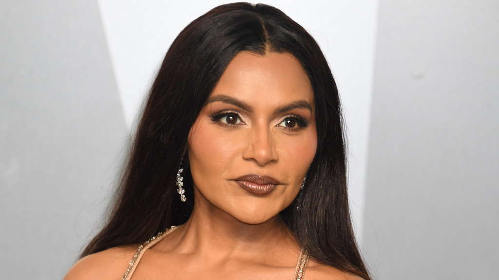 Mindy Kaling looks forward with long dark hair parted in the middle, wearing dark eye makeup, glossy dark lipstick, and sparkling drop earrings.