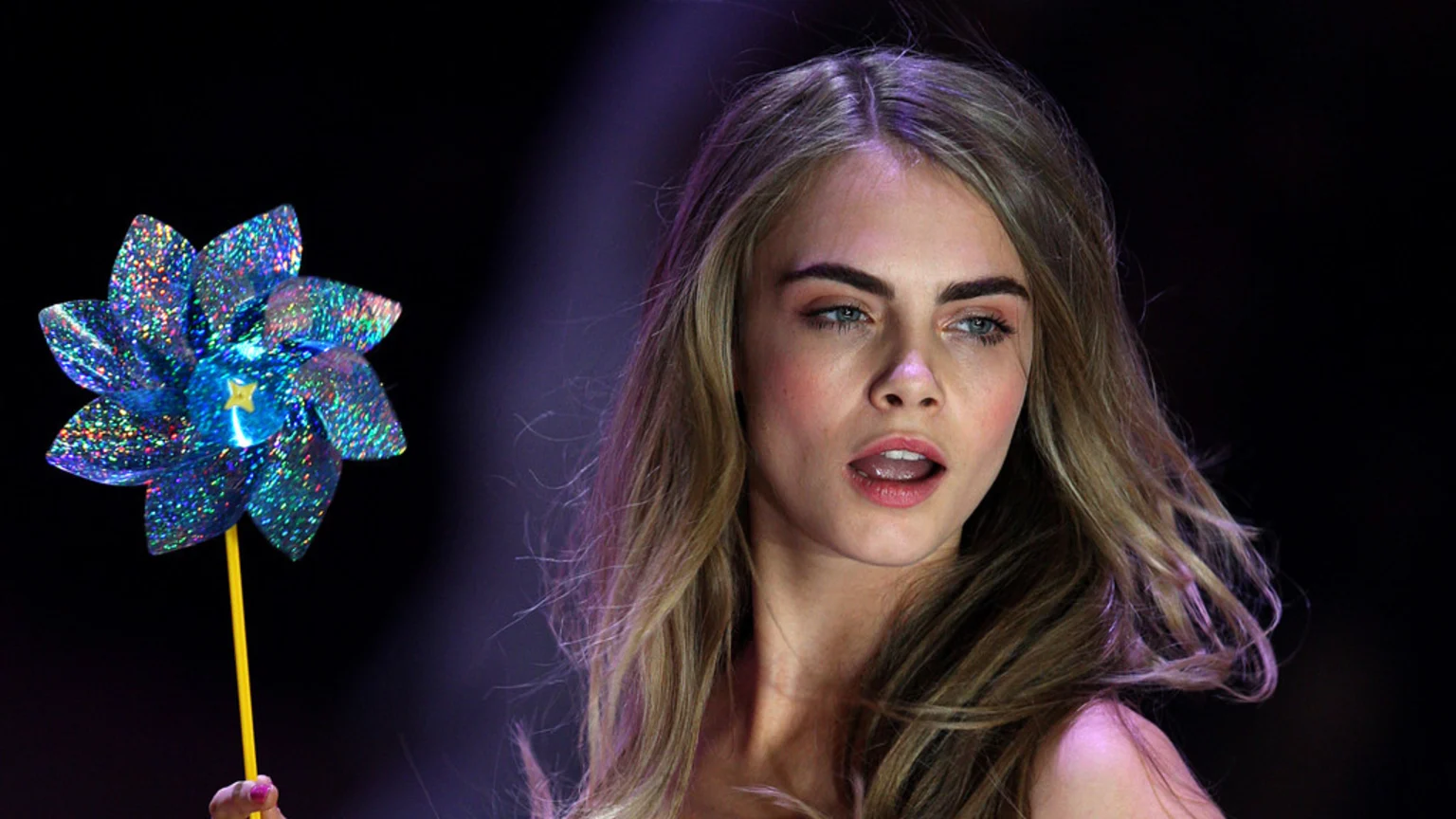 Cara Delevingne, a woman with long blonde hair, looks to the side with her mouth open, holding a blue glittery pinwheel against a dark background.