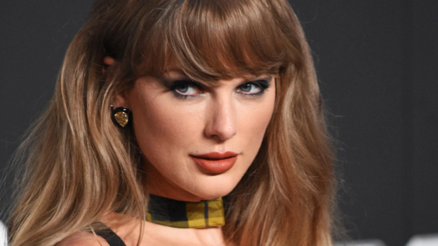 Close-up of Taylor Swift with long blonde hair and bangs, wearing dark eye makeup, reddish lipstick, a plaid choker, and an ornate earring, looking intently at the camera.