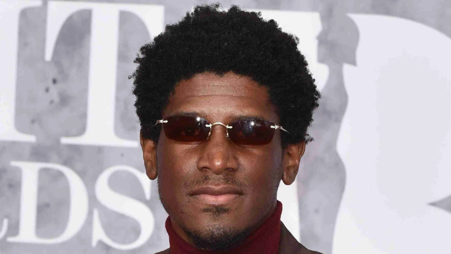 A man with dark skin, an afro, and a goatee, wearing dark rimless sunglasses and a red turtleneck, looks directly at the camera against a blurred grey background.