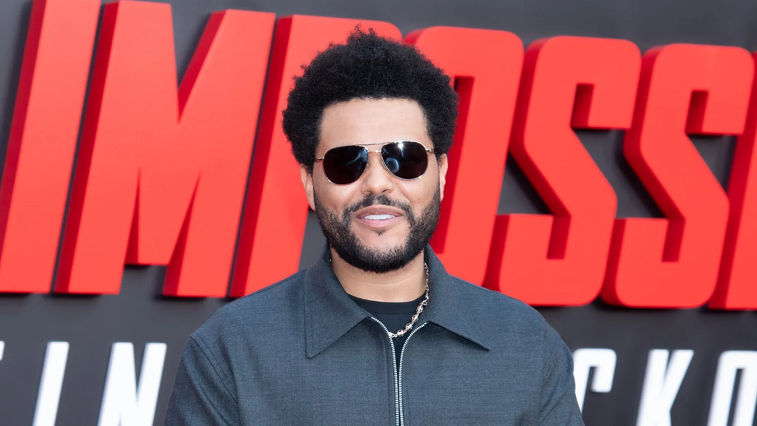 The Weeknd with an afro, beard, and sunglasses, wearing a grey jacket, smiles in front of a large red "IMPOSSIBLE" sign.