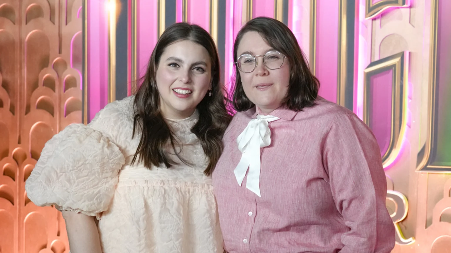 Two smiling women pose against a vibrant pink and gold art deco background; one wears a light dress with textured puff sleeves, the other a pink shirt, white bow, and glasses.