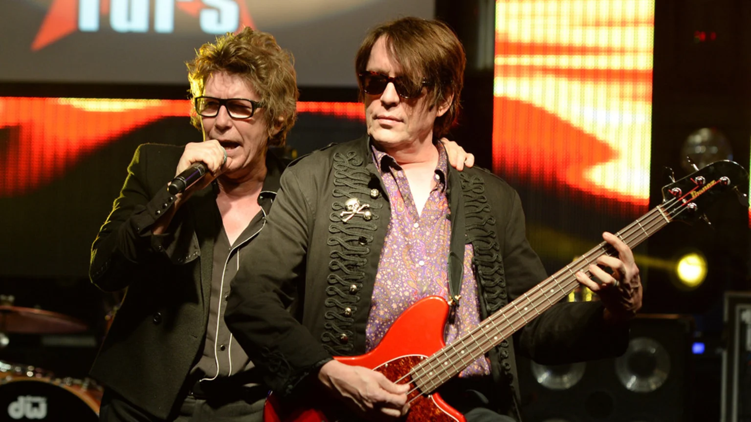 Two musicians perform on stage; one sings into a microphone, wearing glasses, and the other plays a red bass guitar, wearing sunglasses and a military-style jacket with a skull pin.