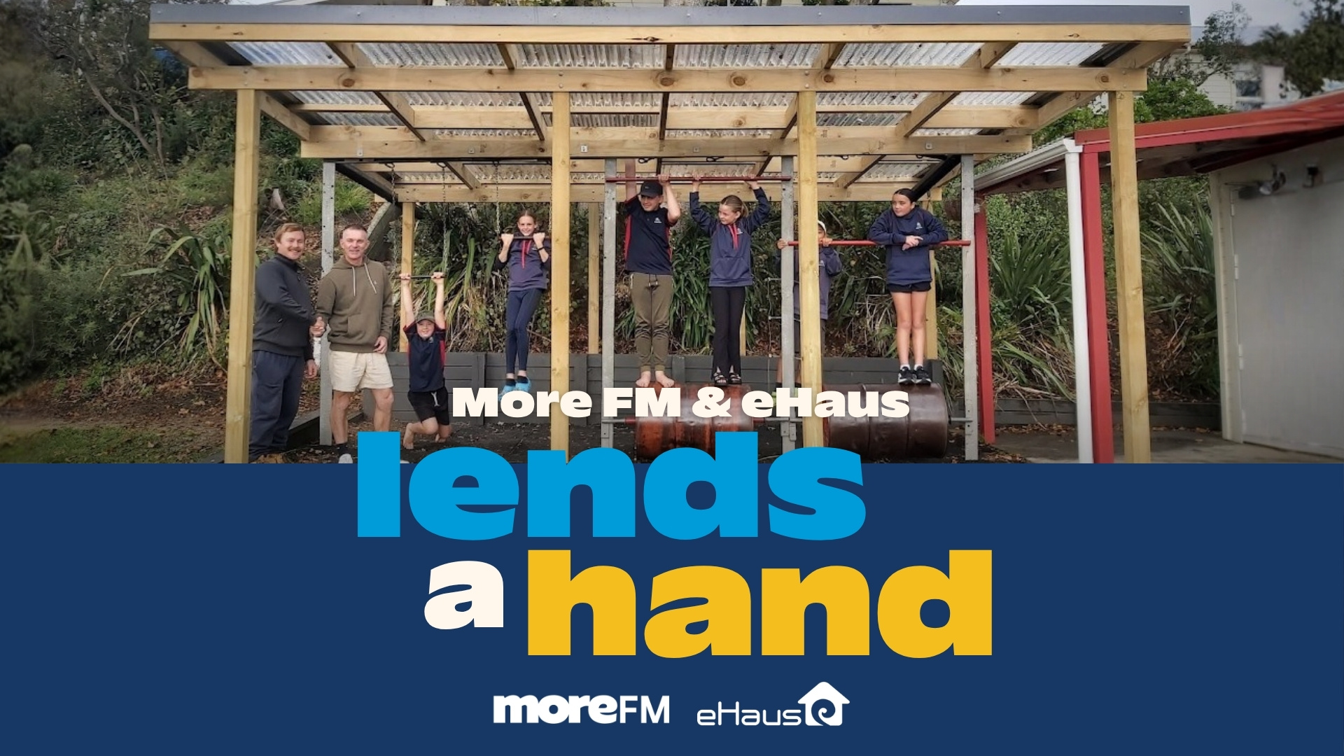 A group of adults and children are gathered around a wooden outdoor exercise structure, with two adults shaking hands and children using the bars, above a banner reading 'More FM & eHaus lends a hand'.