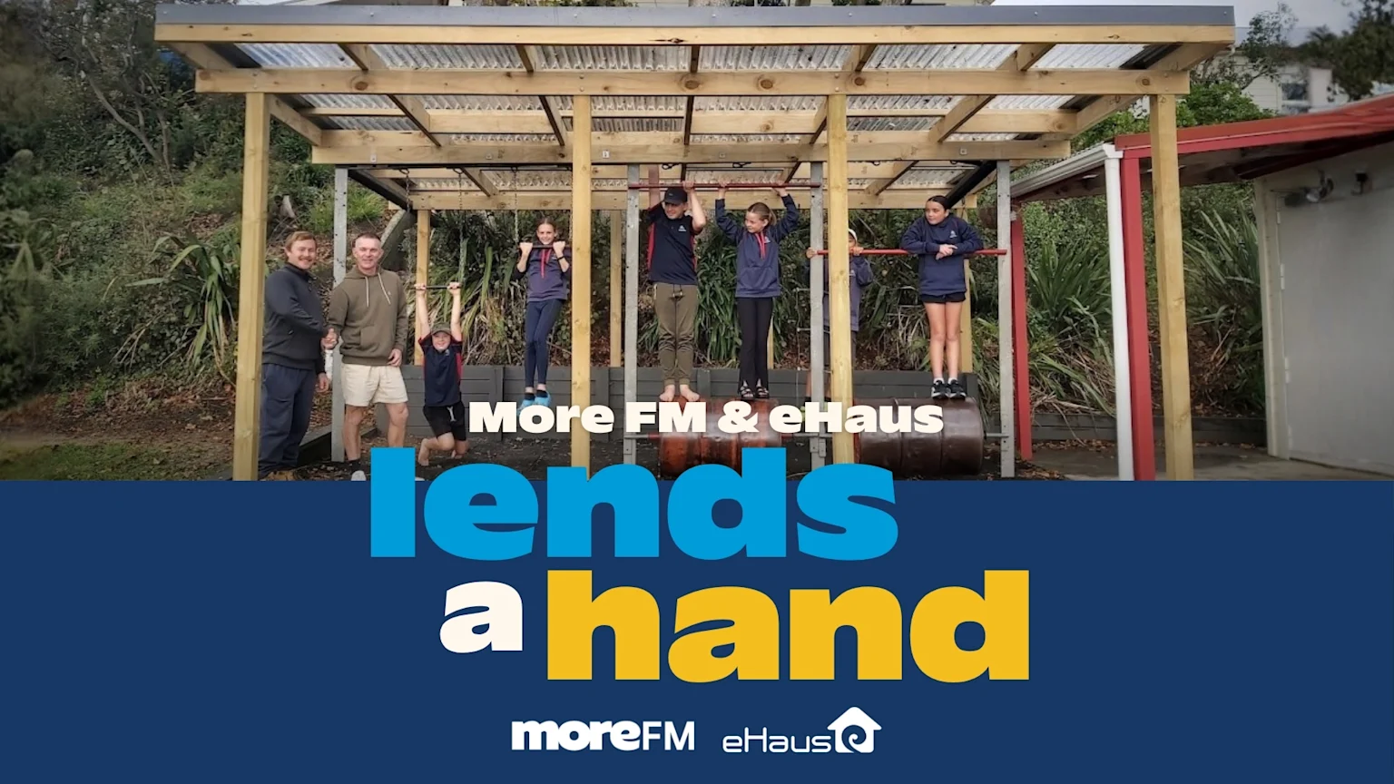 A group of adults and children are gathered around a wooden outdoor exercise structure, with two adults shaking hands and children using the bars, above a banner reading 'More FM & eHaus lends a hand'.
