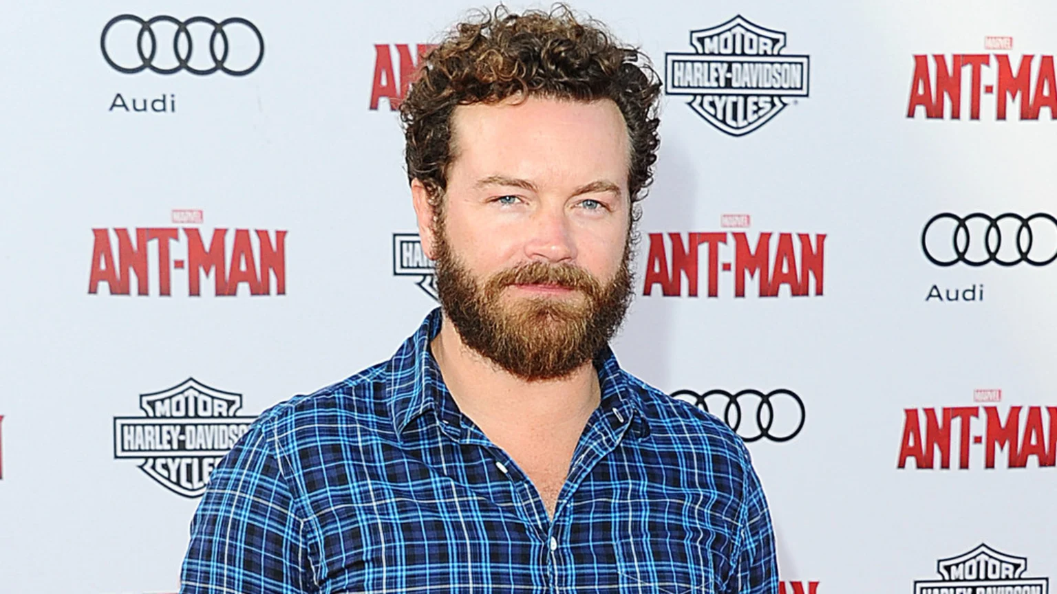 Danny Masterson, a man with curly brown hair and a beard, wears a blue plaid shirt and stands before a backdrop with "Ant-Man," Audi, and Harley-Davidson logos.