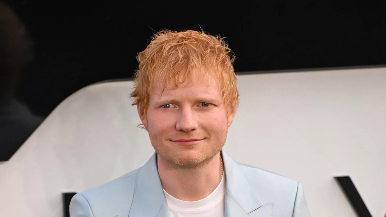 Close-up of Ed Sheeran, a man with red hair and a light beard, smiling slightly at the camera while wearing a light blue blazer over a white t-shirt, against a blurred black and white background.