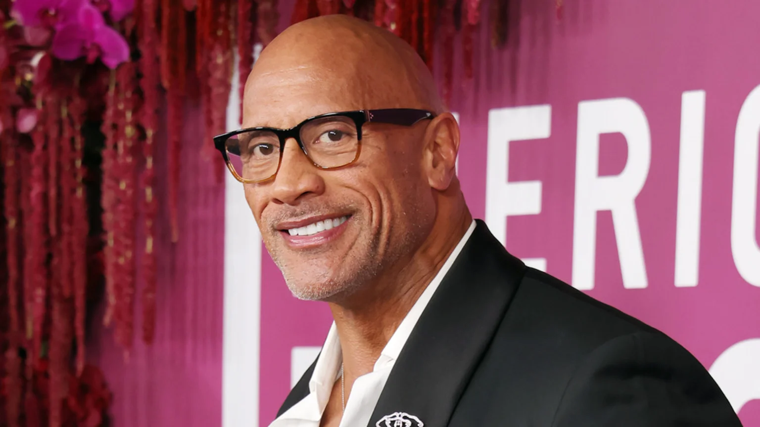 Dwayne "The Rock" Johnson, a bald man with glasses and a goatee, smiles while wearing a suit in front of a blurred pink background with decorative elements and some white text.
