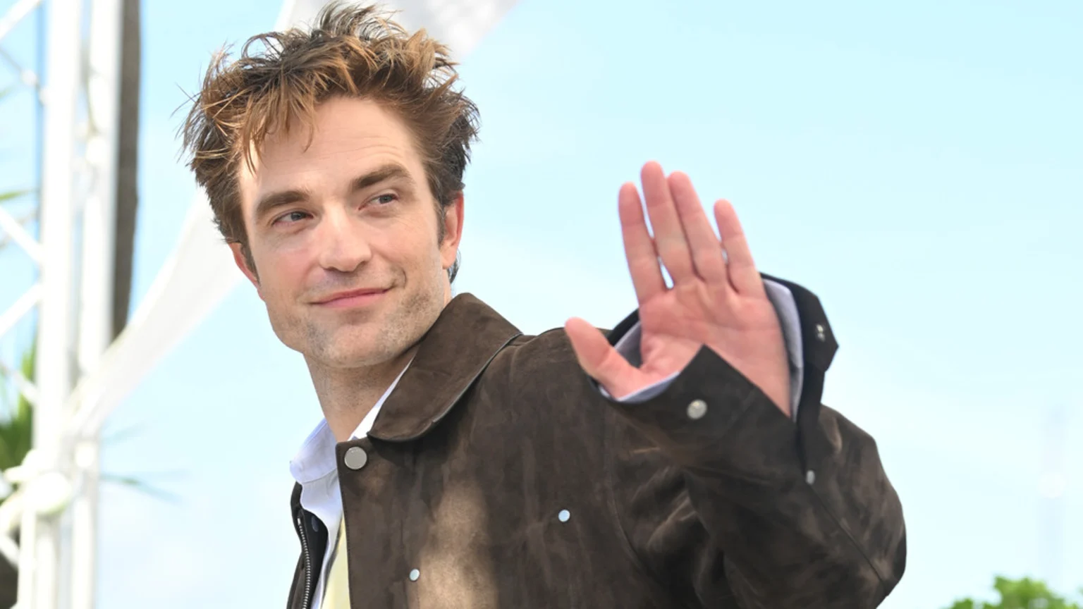 Robert Pattinson smiles, looking to the right, and raises his right hand in a wave, wearing a dark brown jacket against a bright blue sky.