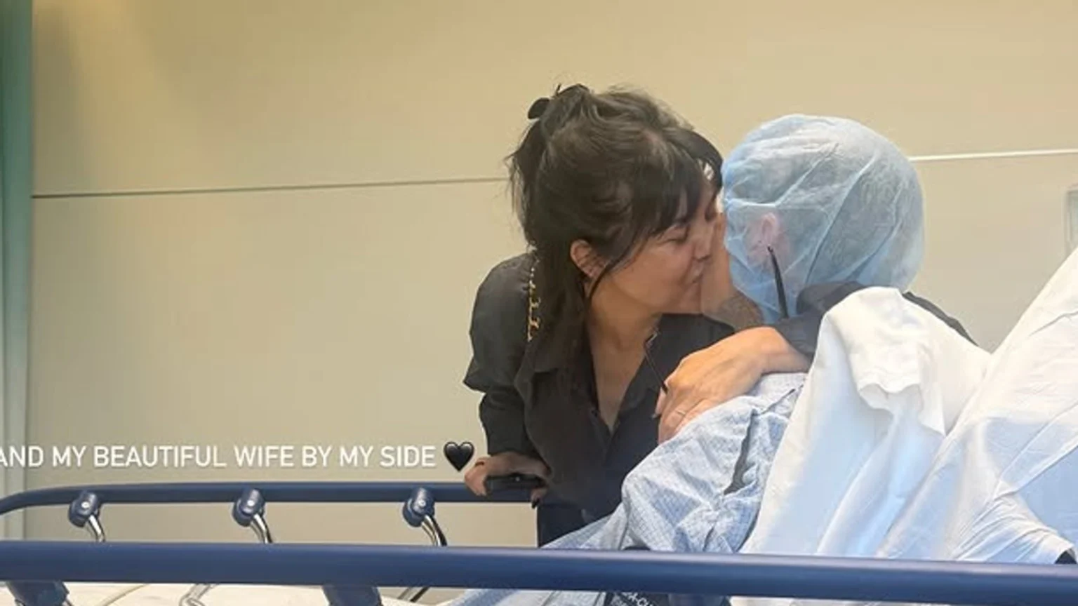 A woman with dark hair leans over to kiss a person wearing a blue surgical cap in a hospital bed, with the text "AND MY BEAUTIFUL WIFE BY MY SIDE ❤️" visible in the bottom left.