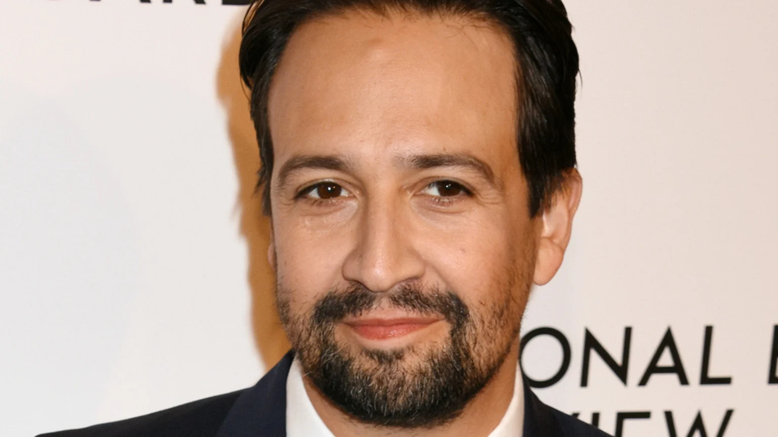 Close-up portrait of Lin-Manuel Miranda smiling subtly, wearing a dark suit jacket and white shirt against a light background with faint text.