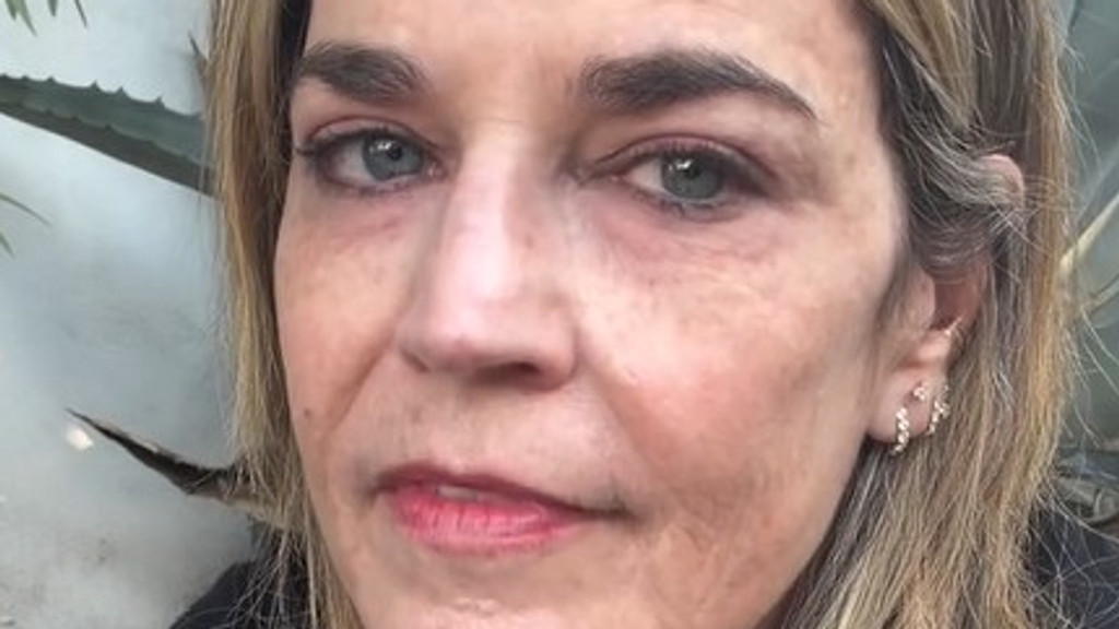 A close-up portrait of a middle-aged woman with blonde hair and blue eyes, looking directly at the camera with a neutral expression. Her face shows natural skin texture with some fine lines, and she wears light makeup and delicate sparkling earrings. A blurred green plant with spiky leaves is visible in the background.