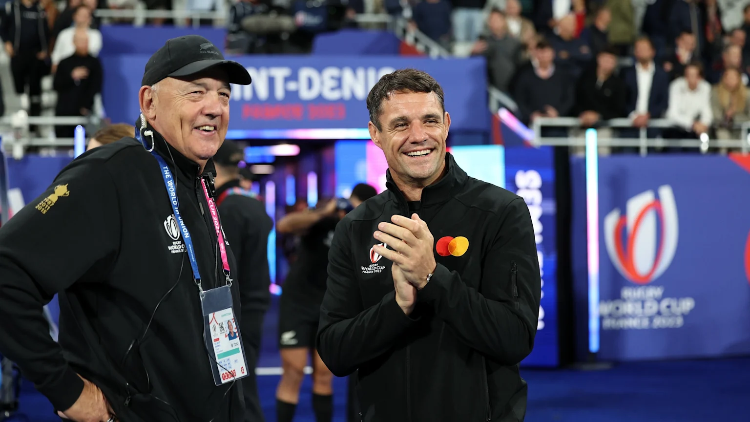 Gilbert Enoka in a black cap and jacket smiles next to a clapping Dan Carter, both at the Rugby World Cup France 2023 event.