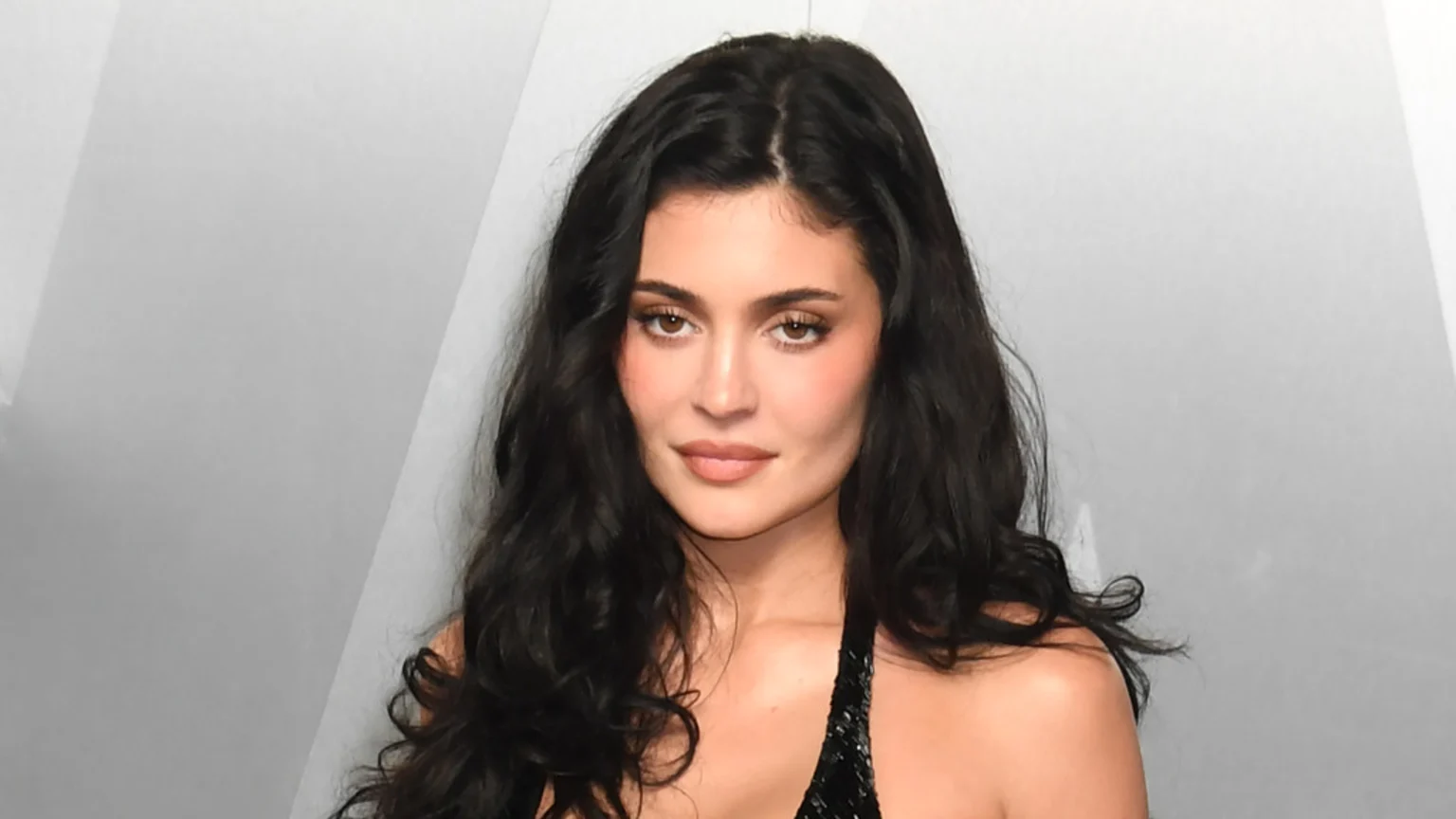 Close-up shot of Kylie Jenner with dark, wavy hair and a neutral expression, looking directly at the camera, wearing a dark top against a light gray background.