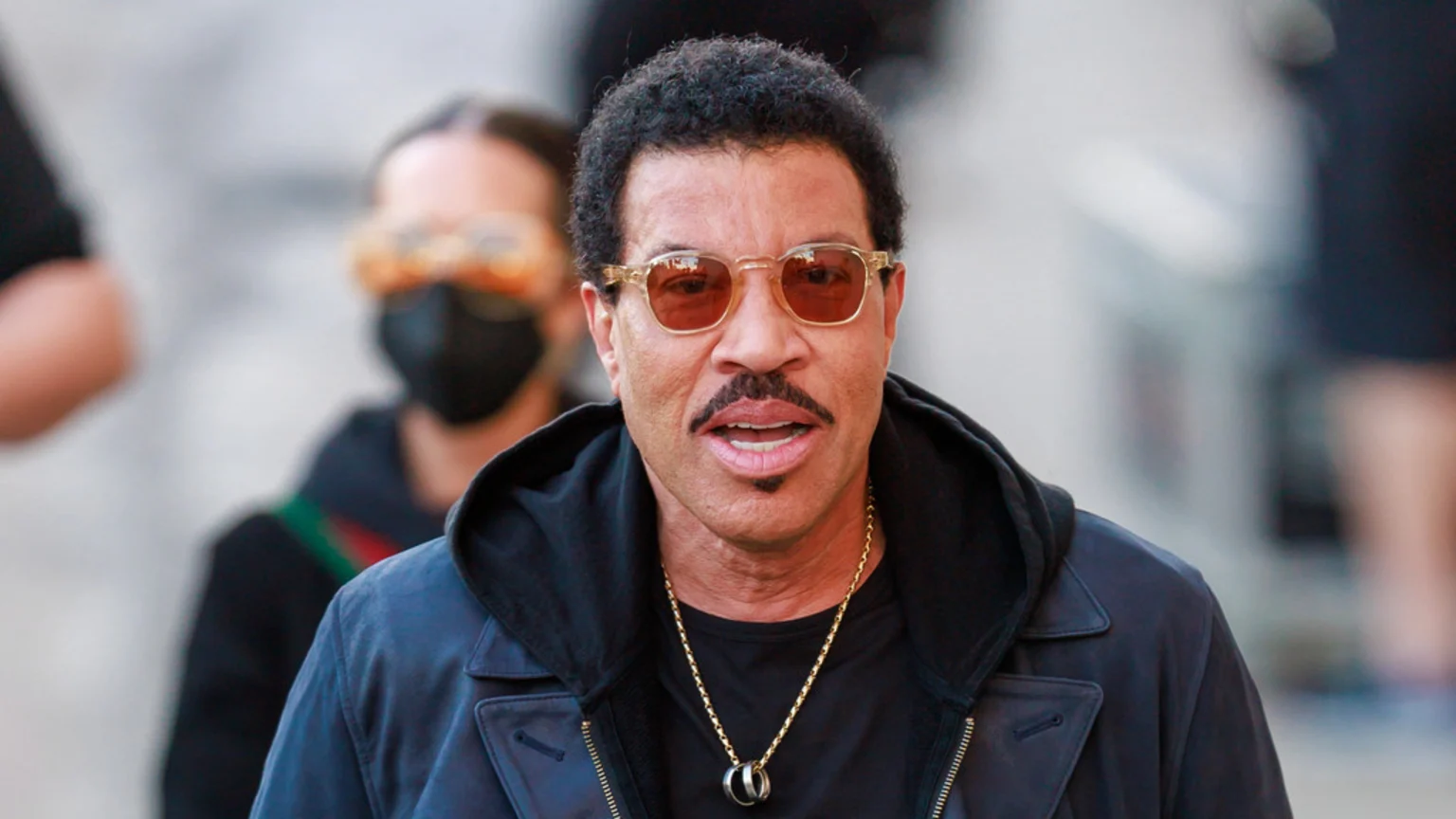 A close-up of Lionel Richie, wearing sunglasses, a dark jacket, and a gold chain with a pendant, looking directly at the viewer, with a blurry figure behind him.