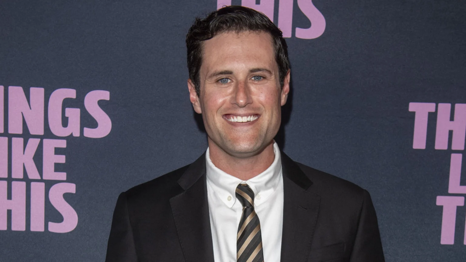 A smiling man with dark hair and blue eyes wears a black suit, white shirt, and striped tie against a dark background with light purple text partly reading "INGS LIKE HIS" and "TH".