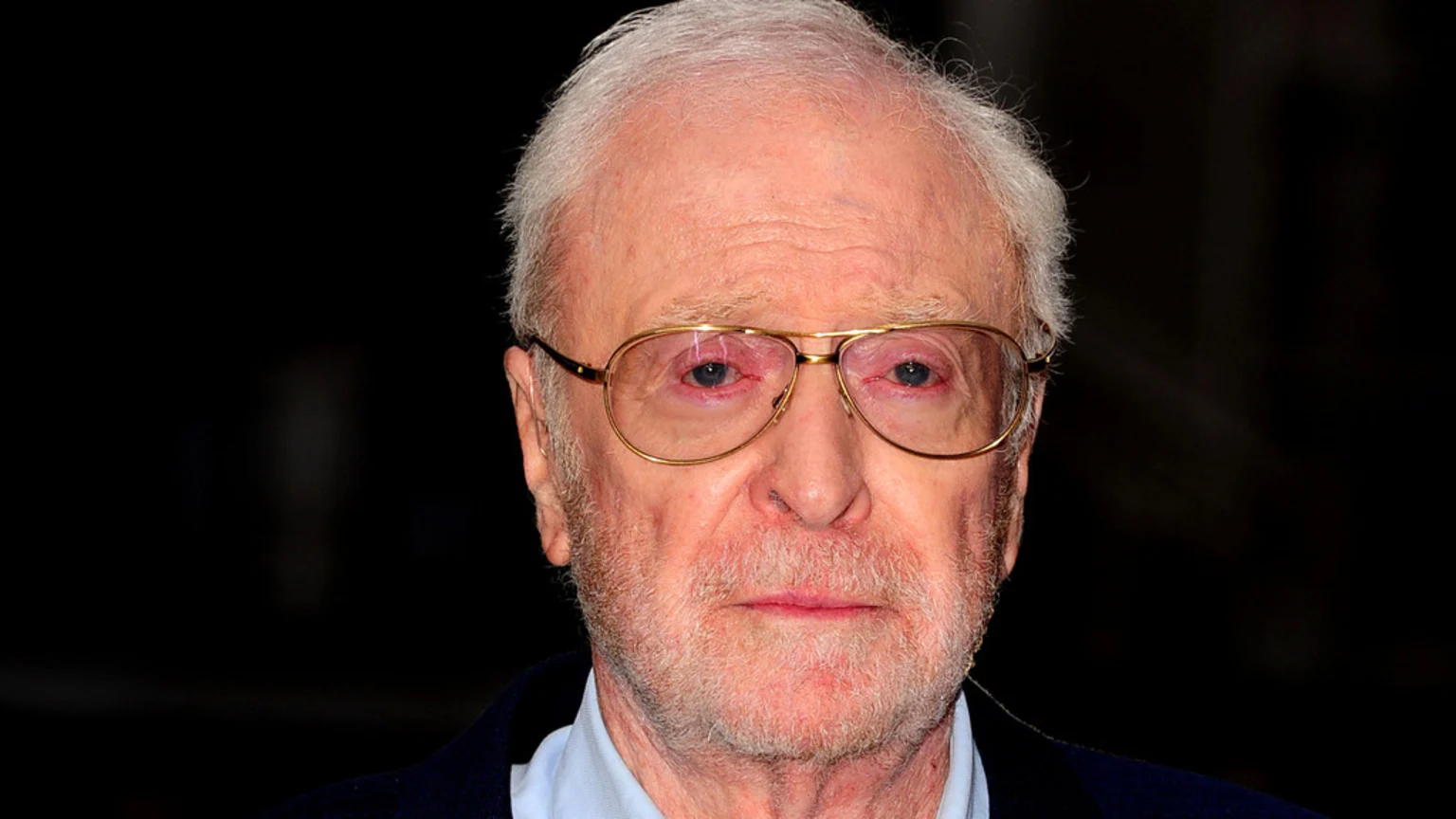 Close-up of actor Michael Caine, an elderly man with white hair and beard, wearing gold-rimmed glasses and a blue shirt, looking directly ahead with red eyes.