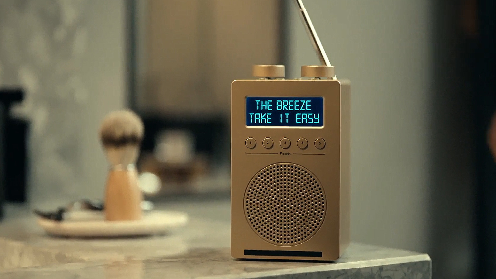 A gold radio with an extended antenna displays "THE BREEZE" and "TAKE IT EASY" on its blue screen, on a counter with a blurry shaving brush and razor.