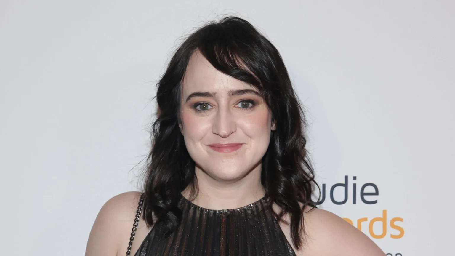 A woman with dark, wavy hair and bangs smiles faintly at the camera, wearing a dark pleated top against a white background with a partial "Audie Awards" logo.
