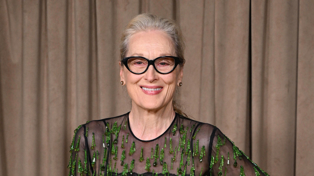 Meryl Streep smiles wearing black cat-eye glasses, a sheer black top with green embellishments, and small earrings against a brown curtain background.