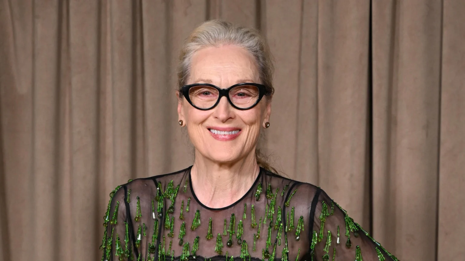 Meryl Streep smiles wearing black cat-eye glasses, a sheer black top with green embellishments, and small earrings against a brown curtain background.