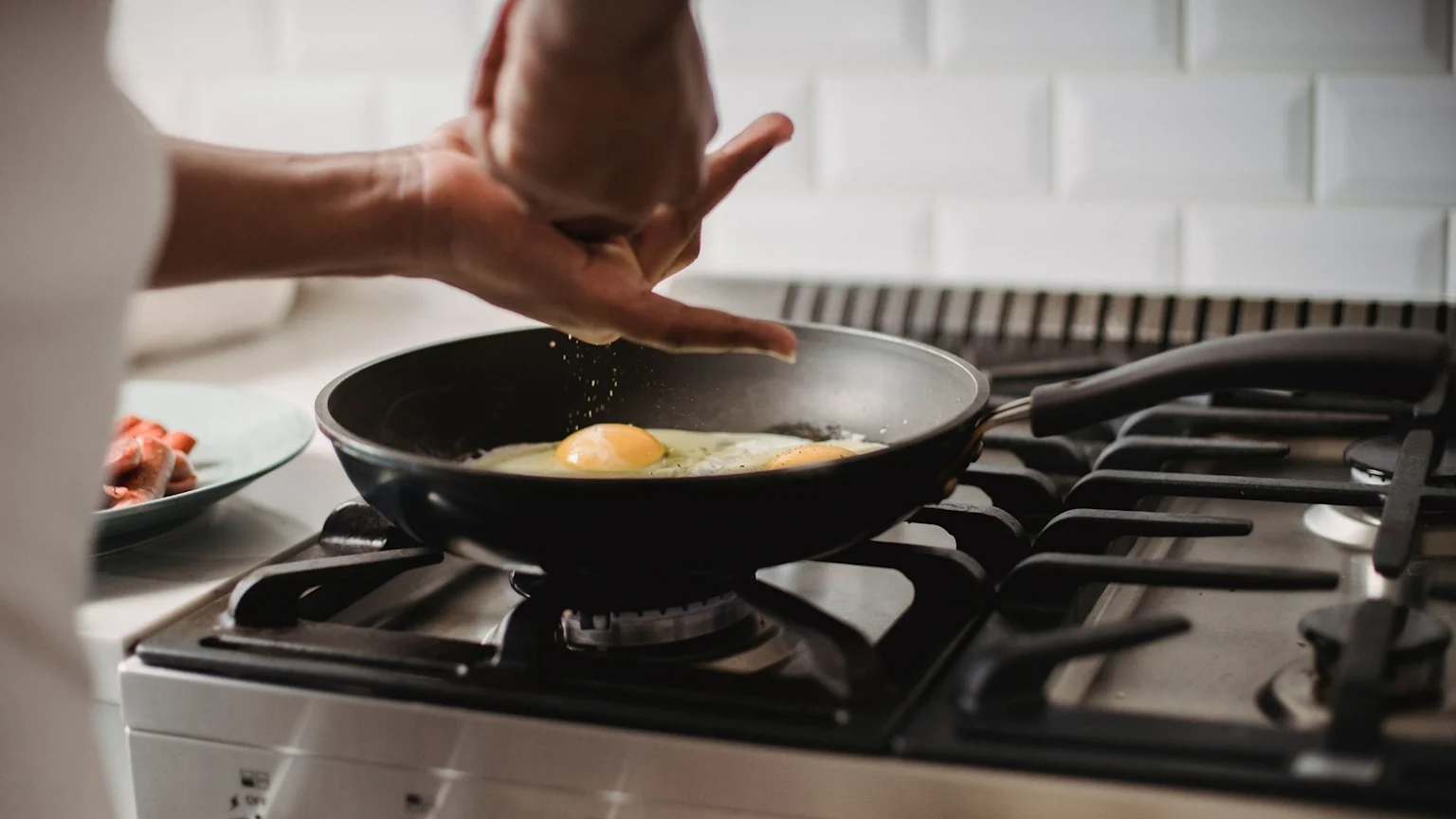 A person's hand sprinkles seasoning over two frying eggs in a pan on a gas stove, with a plate of cooked sausages visible on the side.