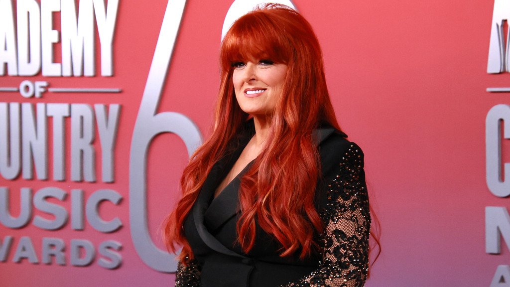 Wynonna Judd, with long red hair, smiles wearing a black top with sequined lace sleeves, standing against a red background featuring "ACADEMY OF COUNTRY MUSIC AWARDS" text.