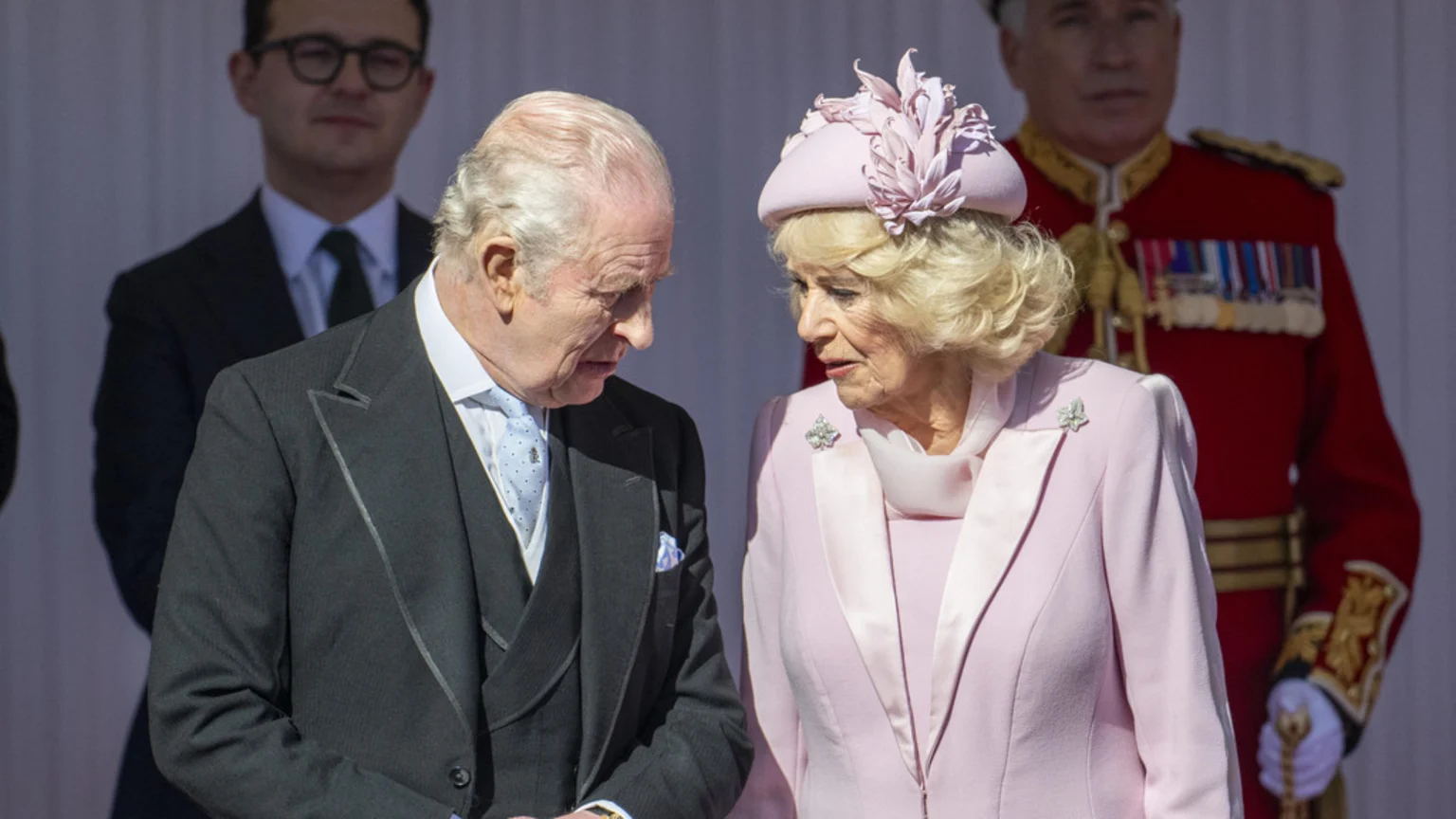 King Charles III in a dark suit and Queen Camilla in a pink suit and hat stand facing each other, appearing to converse, with two blurred men behind them.