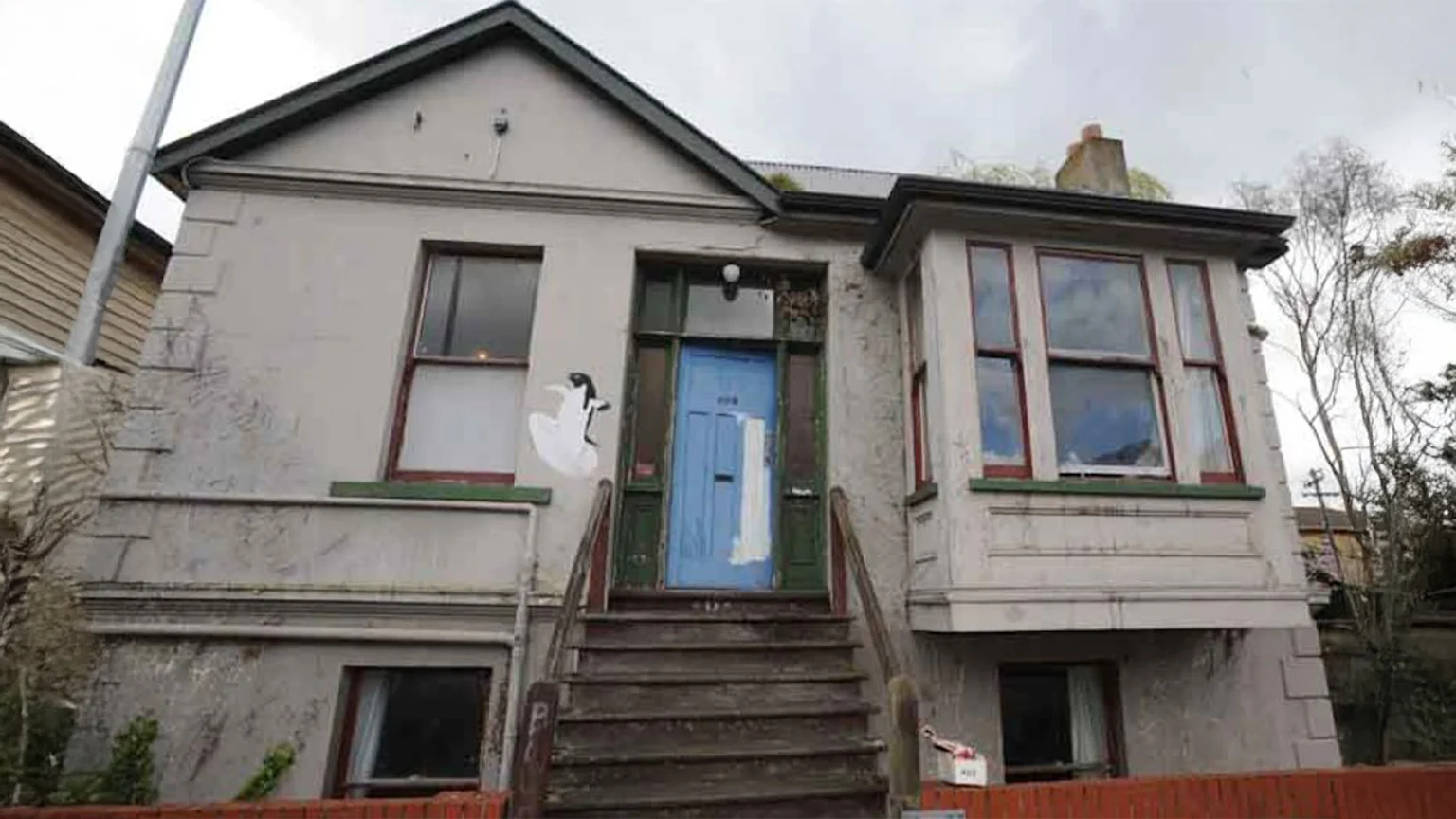 The front exterior of a somewhat dilapidated light-colored house or flat with a blue front door, wooden steps, red-framed windows, and a white bird stencil on the wall.