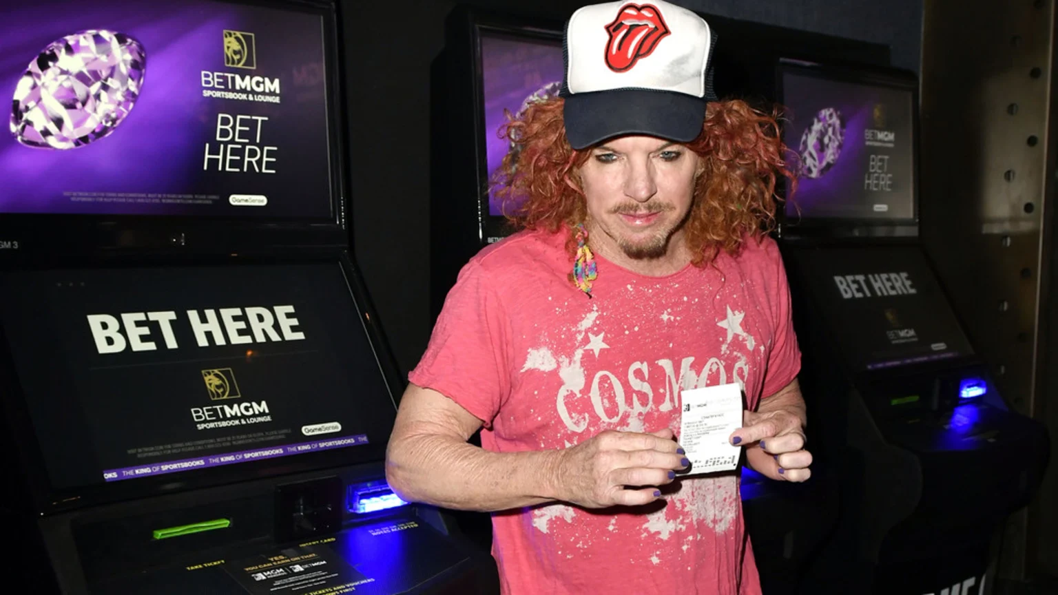 Comedian Carrot Top, wearing a Rolling Stones hat, pink t-shirt, and purple nail polish, holds a ticket in front of multiple BetMGM betting kiosks.