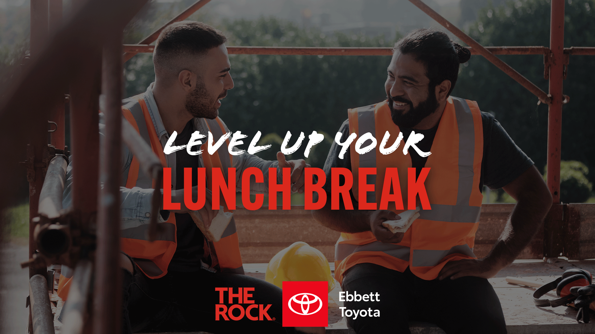 Two smiling construction workers in high-vis vests eat and talk during a lunch break, with "LEVEL UP YOUR LUNCH BREAK" and "THE ROCK. Ebbett Toyota" branding.