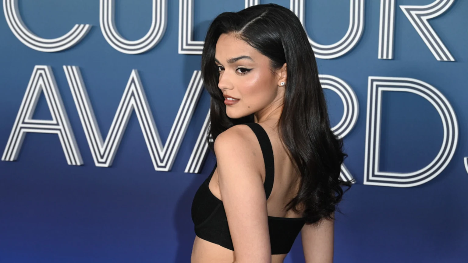 A woman with long dark hair and winged eyeliner looks over her right shoulder, wearing a black top, against a blue background with white striped letters spelling "CULTURE AWARDS."