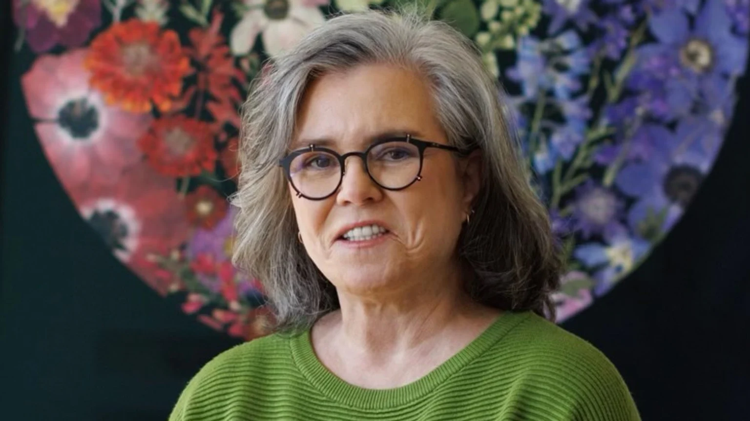 A woman with gray hair and glasses, wearing a green ribbed sweater, looks forward with a tense expression in front of a colorful, blurred floral background.