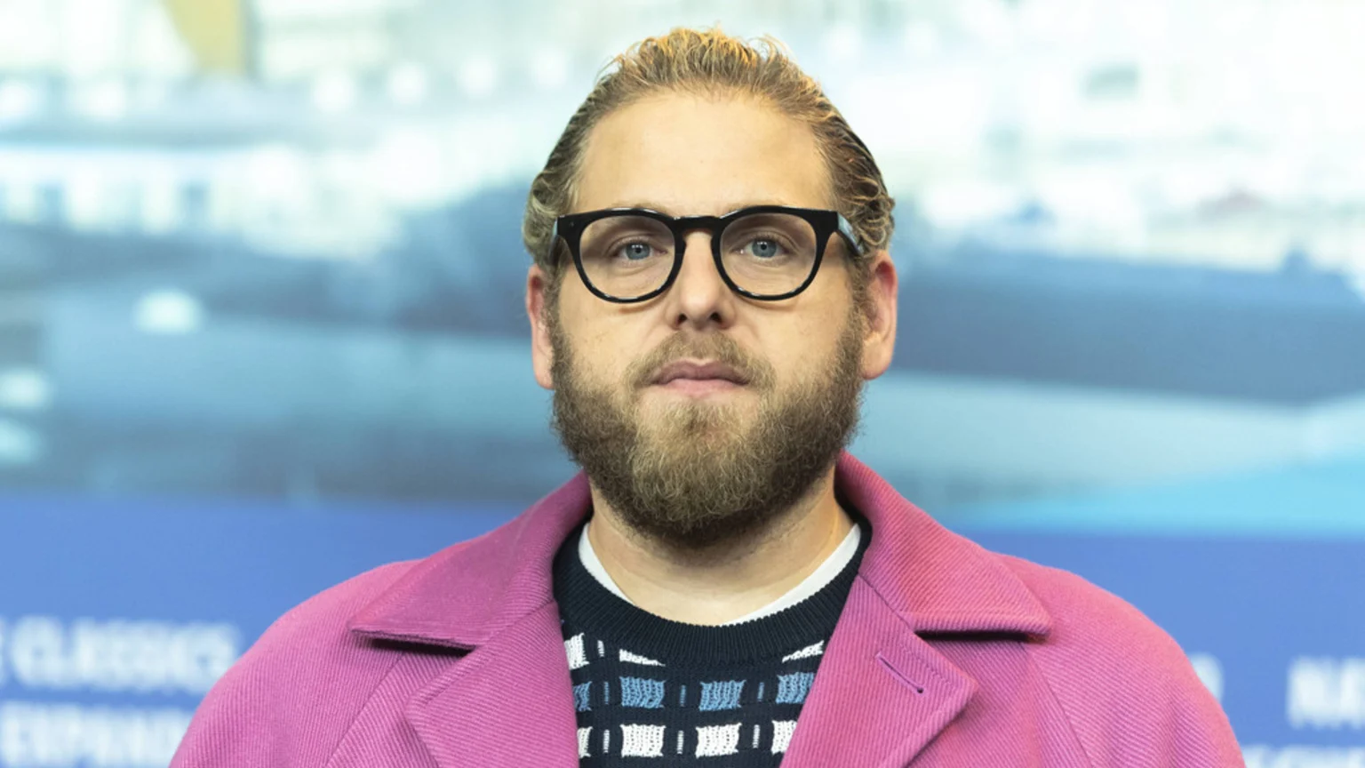 Jonah Hill with a beard and glasses, wearing a pink coat over a patterned sweater, looking forward against a blurred blue background.