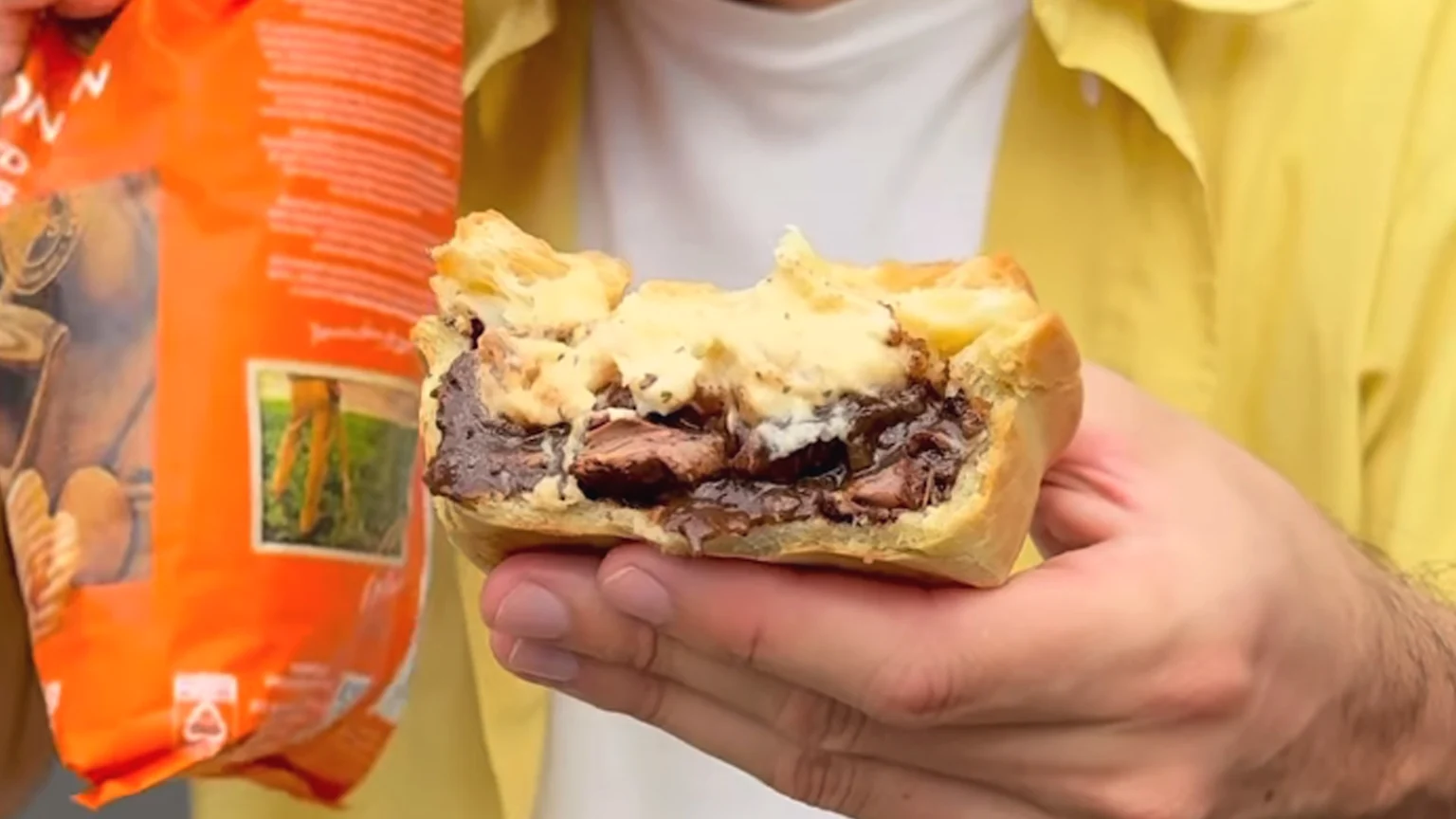A person holds a golden-crusted savory pie, bitten to show a dark meat filling and a creamy mashed potato or cheese topping, with an orange package in the background.