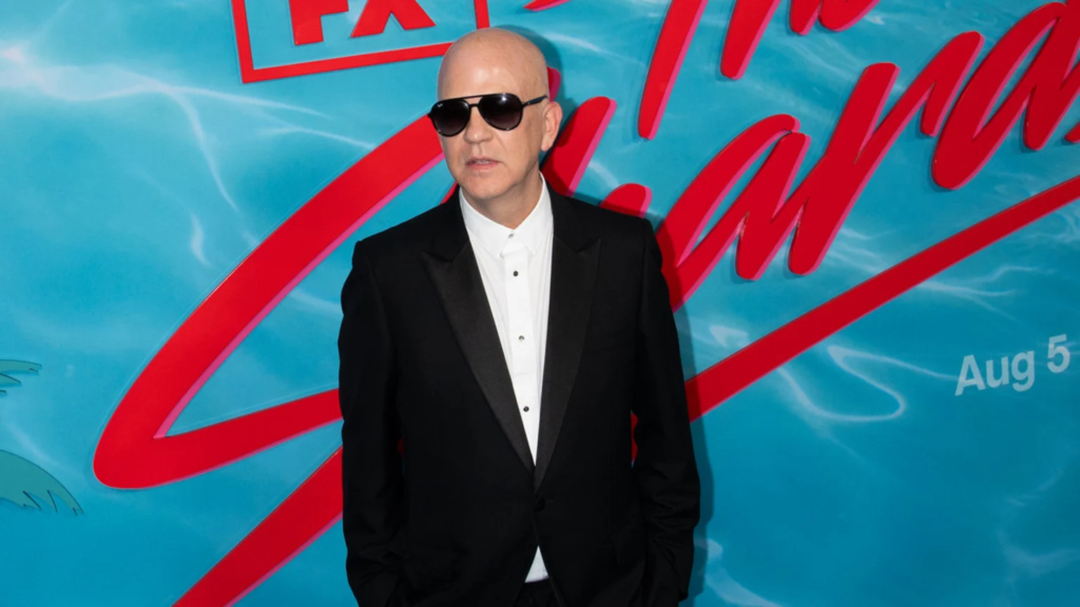 A bald man in sunglasses, wearing a black tuxedo and white shirt, stands against a bright blue backdrop featuring red stylized text and the date 'Aug 5'.