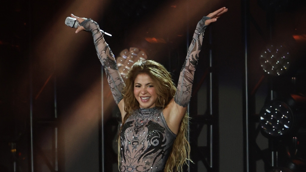 Shakira smiles broadly on stage with her arms raised, wearing a sparkly mesh top and holding a microphone.