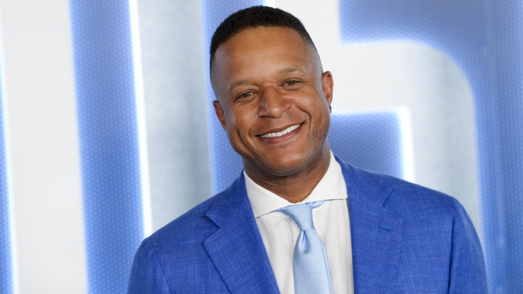 A smiling Black man, Craig Melvin, in a blue suit and light blue tie, poses against a light blue and white striped background.