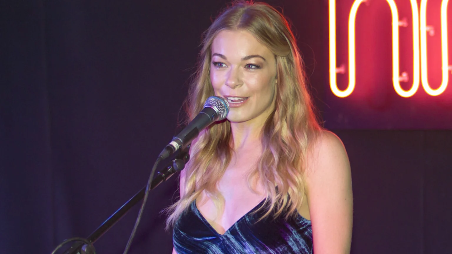 A smiling woman with long blonde hair and a dark blue v-neck top stands at a microphone, with a glowing red neon sign in the blurred background.