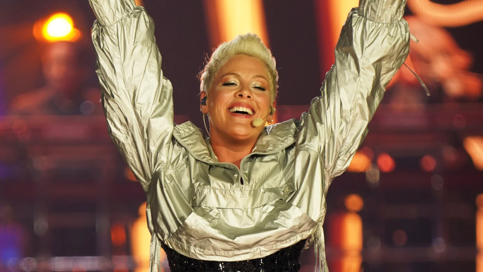 Singer Pink, with short blonde hair, smiles broadly and raises her arms overhead, wearing a shiny silver jacket and a microphone, performing on a stage with blurred warm lights in the background.